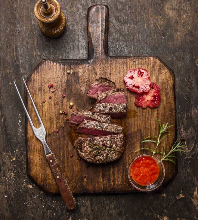 44560560 - roasted ribeye steak sliced on a cutting board with a fork, red sauce, peppers and tomatoes on rustic wooden background, top view